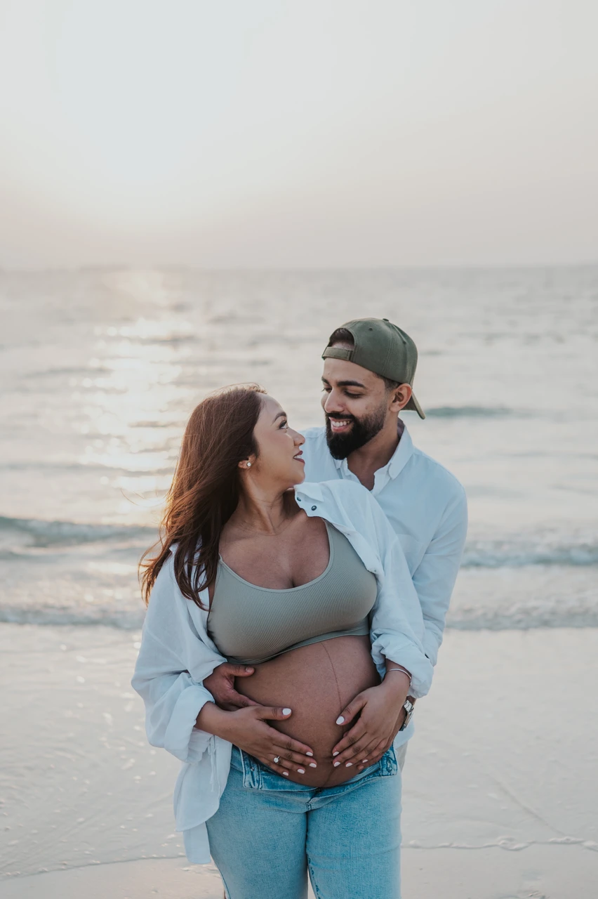 Maternity Shoot Outfit Dubai - casual denim beach set A couple embraces at the Dubai shoreline during a maternity shoot, styled in casual denim and open white layers for a relaxed beach outfit feel.
