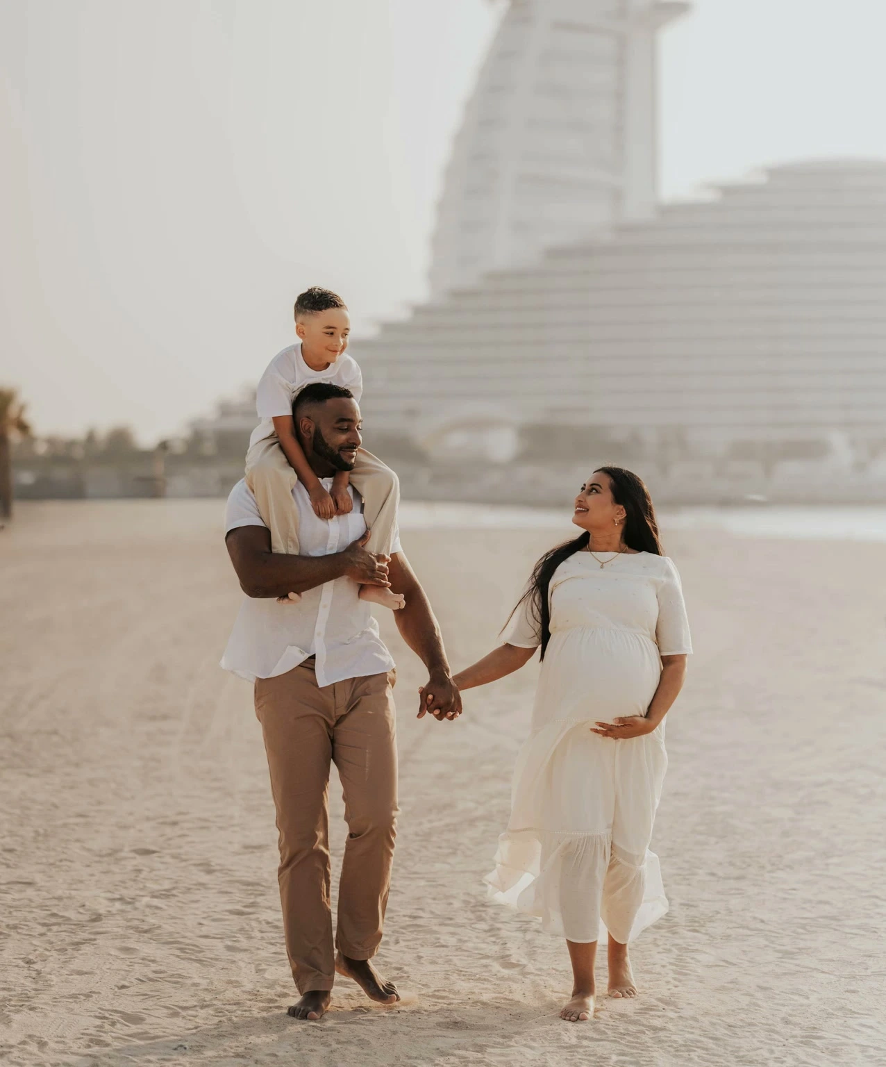 A pregnant mother, father, and child walk on Dubai beach during a maternity shoot, wearing coordinated neutral outfits with Burj Al Arab visible behind them.