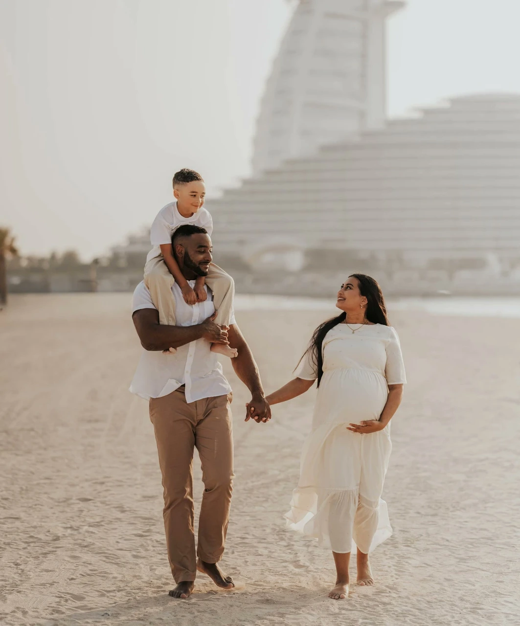 Maternity Shoot Outfit Dubai - Burj beach family neutrals A pregnant mother, father, and child walk on Dubai beach during a maternity shoot, wearing coordinated neutral outfits with Burj Al Arab visible behind them.