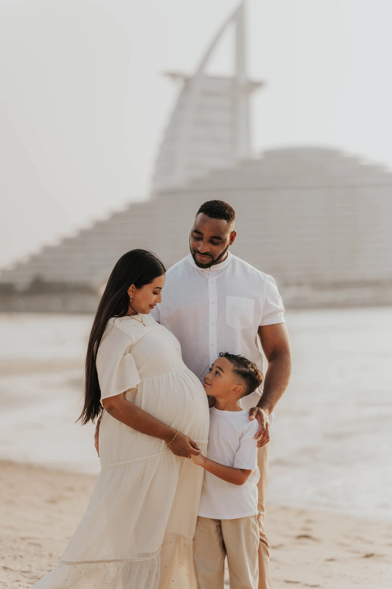 A pregnant mother, father, and son pose on Dubai beach during a maternity shoot, wearing coordinated white and beige outfits for a clean family look.