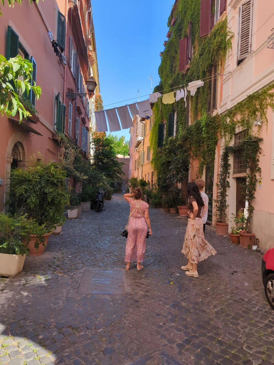 Rome Photoshoot Outfit - Patterned Summer Style Three visitors in patterned summer outfits stand on a cobbled Rome lane beneath hanging laundry and ivy-covered façades.