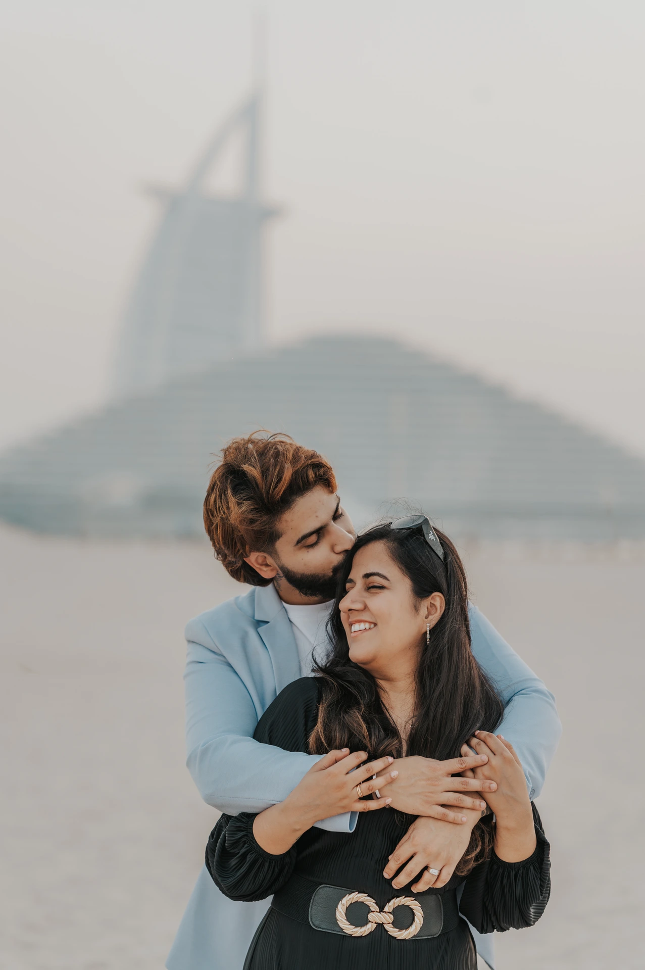 Baiser après la demande près du Burj Al Arab Un homme enlace sa partenaire souriante et l’embrasse sur le front sur la plage, avec le Burj Al Arab derrière leur demande en mariage à Dubaï.