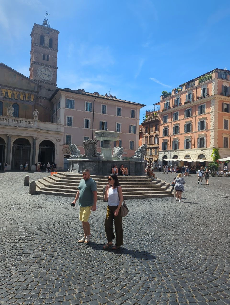 Trastevere Photoshoot - Piazza Santa Maria Visitors cross Piazza di Santa Maria in Trastevere beside its fountain and basilica bell tower under a clear blue sky.
