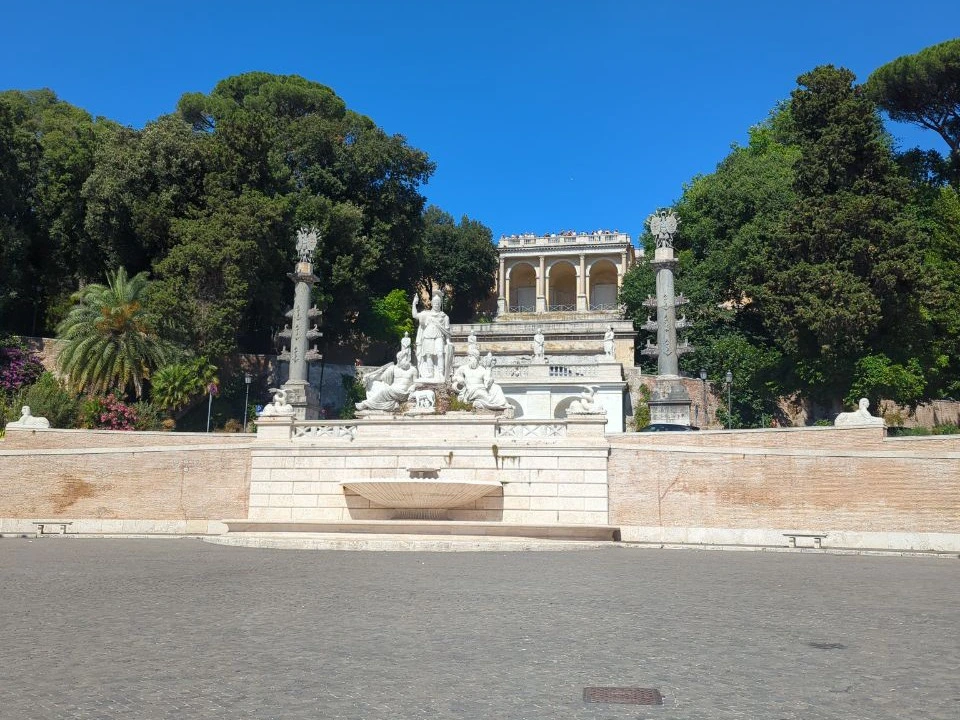 A pale sculptural fountain and paired columns rise beneath the tree-lined Pincio terrace above Piazza del Popolo in Rome.