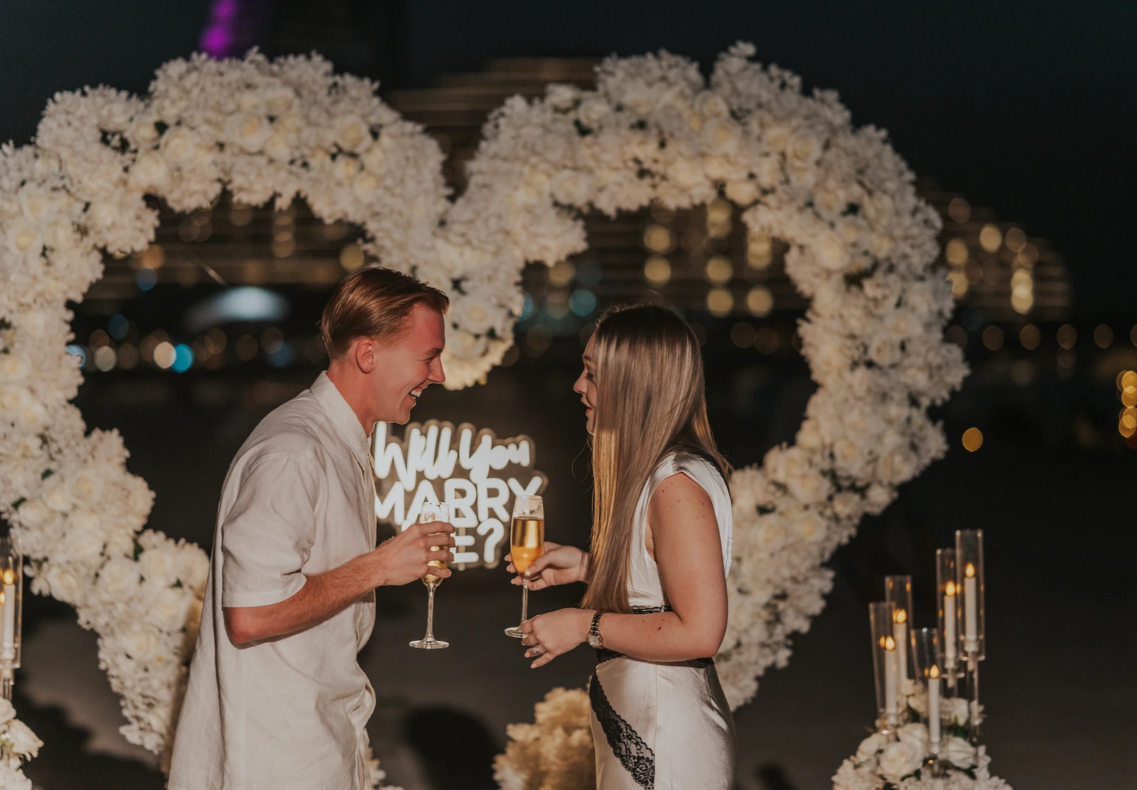 A couple toasts with champagne in front of a white heart proposal setup on Dubai beach at night, celebrating the engagement moment.