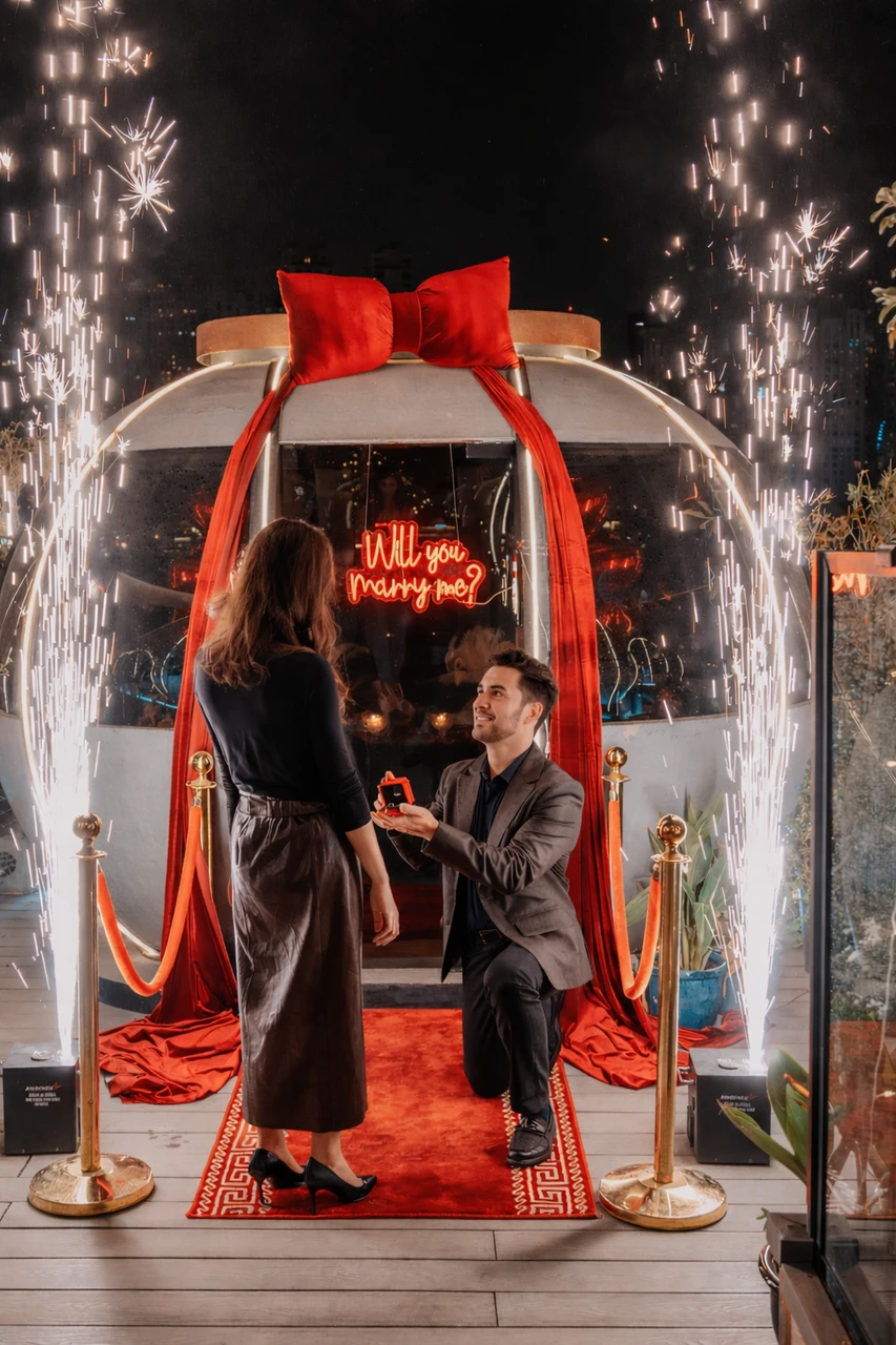 Proposal at the Pods in Dubai - Sparkler surprise proposal A man kneels with a ring box on a red carpet outside The Pods in Dubai, with sparkler fireworks and a “Will you marry me?” sign behind him.
