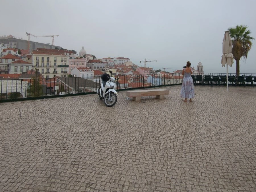 A visitor looks across Alfama’s red roofs from the Portas do Sol terrace beside a scooter, stone bench and black railing.
