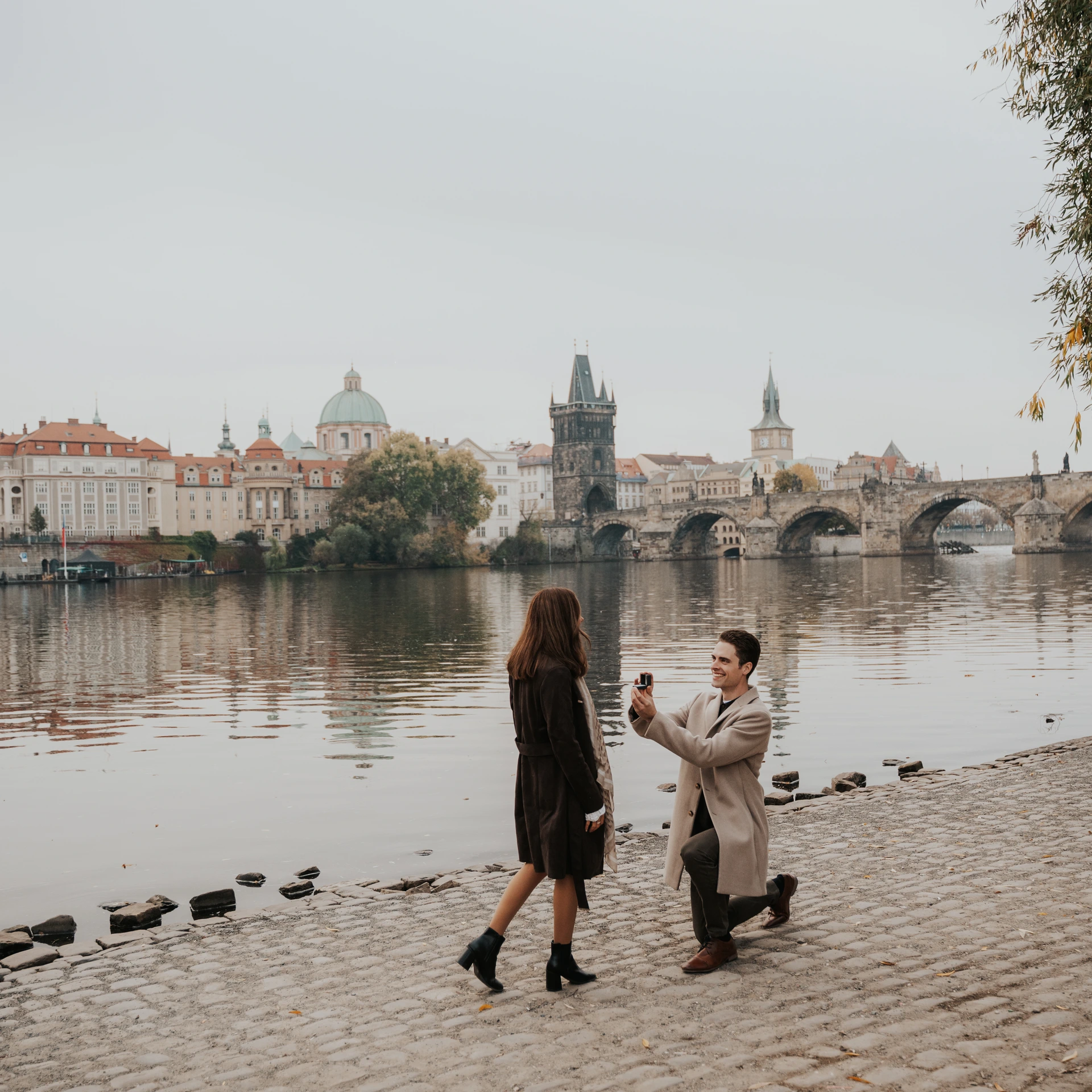 Man kneeling with a ring box in front of his partner beside the Vltava in Prague, with Charles Bridge behind them.