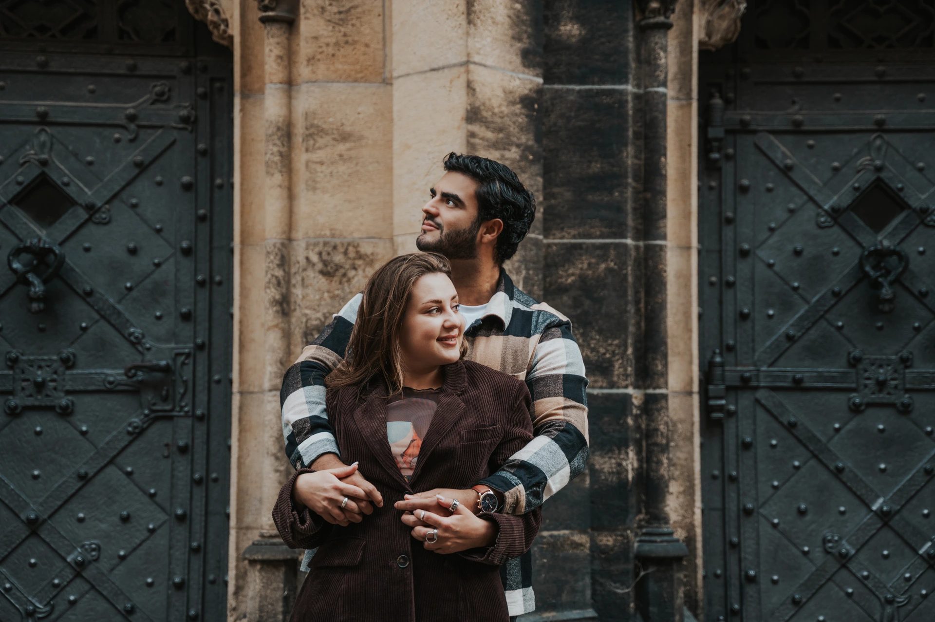 Couple embracing in front of a sandstone column between two dark metal doors with decorative ironwork in Prague.
