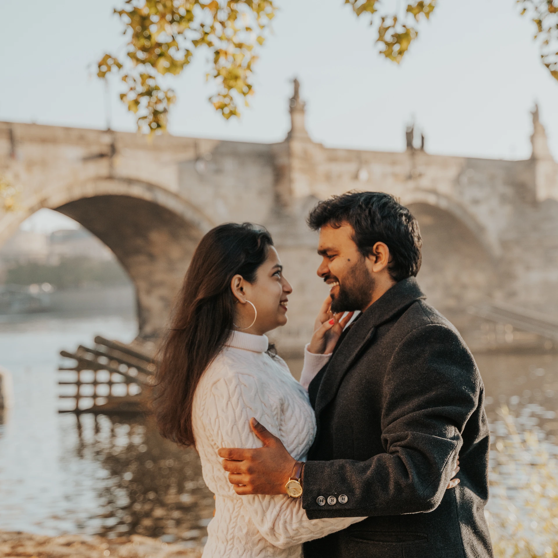 Couple embracing beside the Vltava in Prague, with Charles Bridge and autumn leaves in the background.