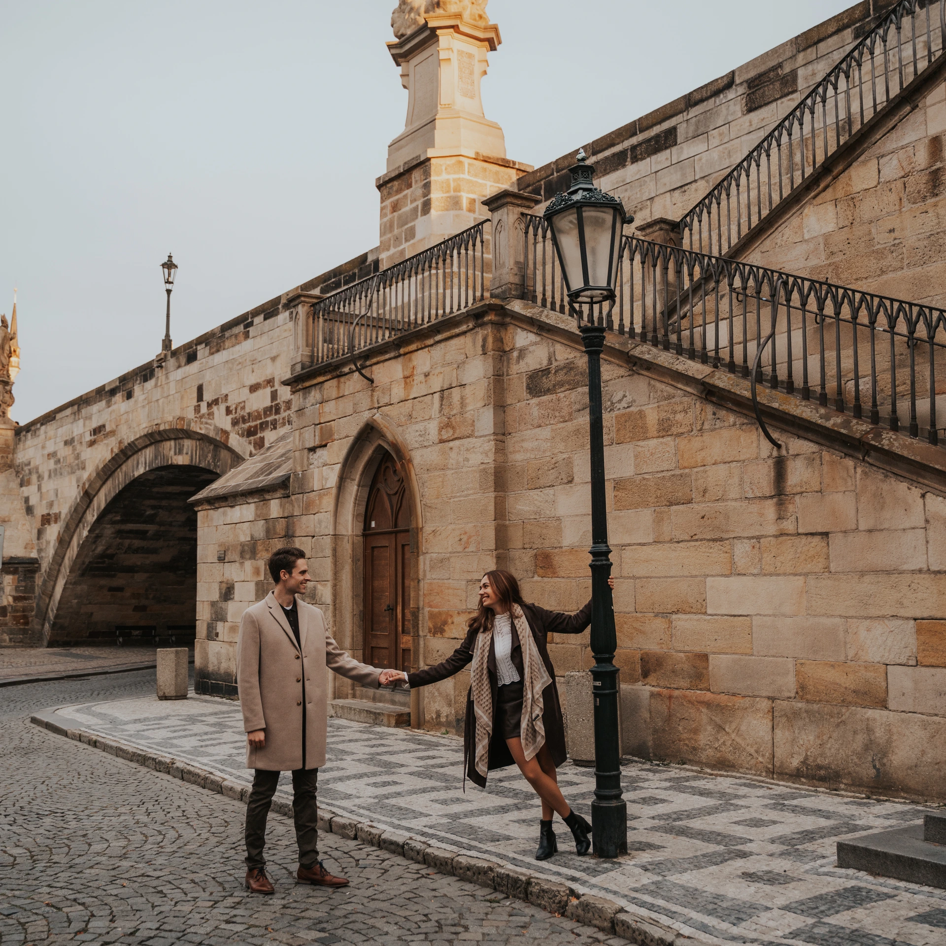 Couple holding hands beside a lamppost below the stone staircase next to Charles Bridge in Prague.