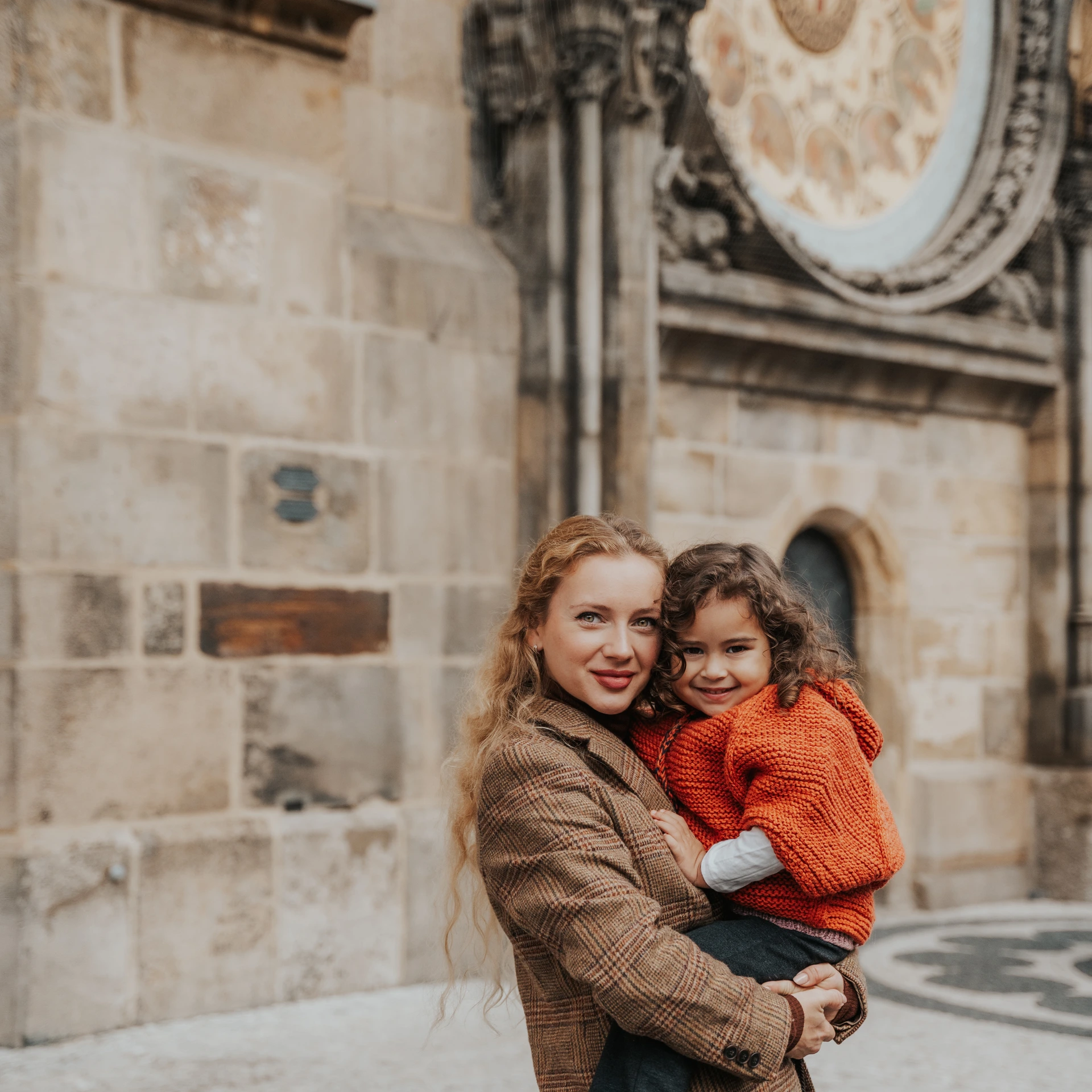 Woman holding a smiling child in an orange jumper in front of the Astronomical Clock in Prague Old Town Square.