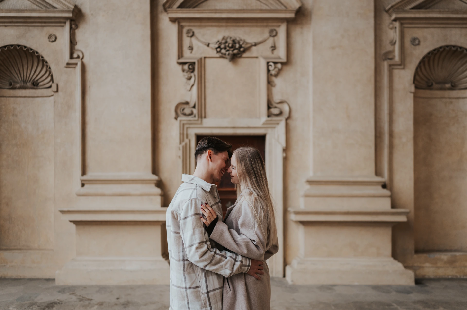 Couple embracing with their foreheads touching in front of a pale stone facade with columns and decorative niches in Prague.