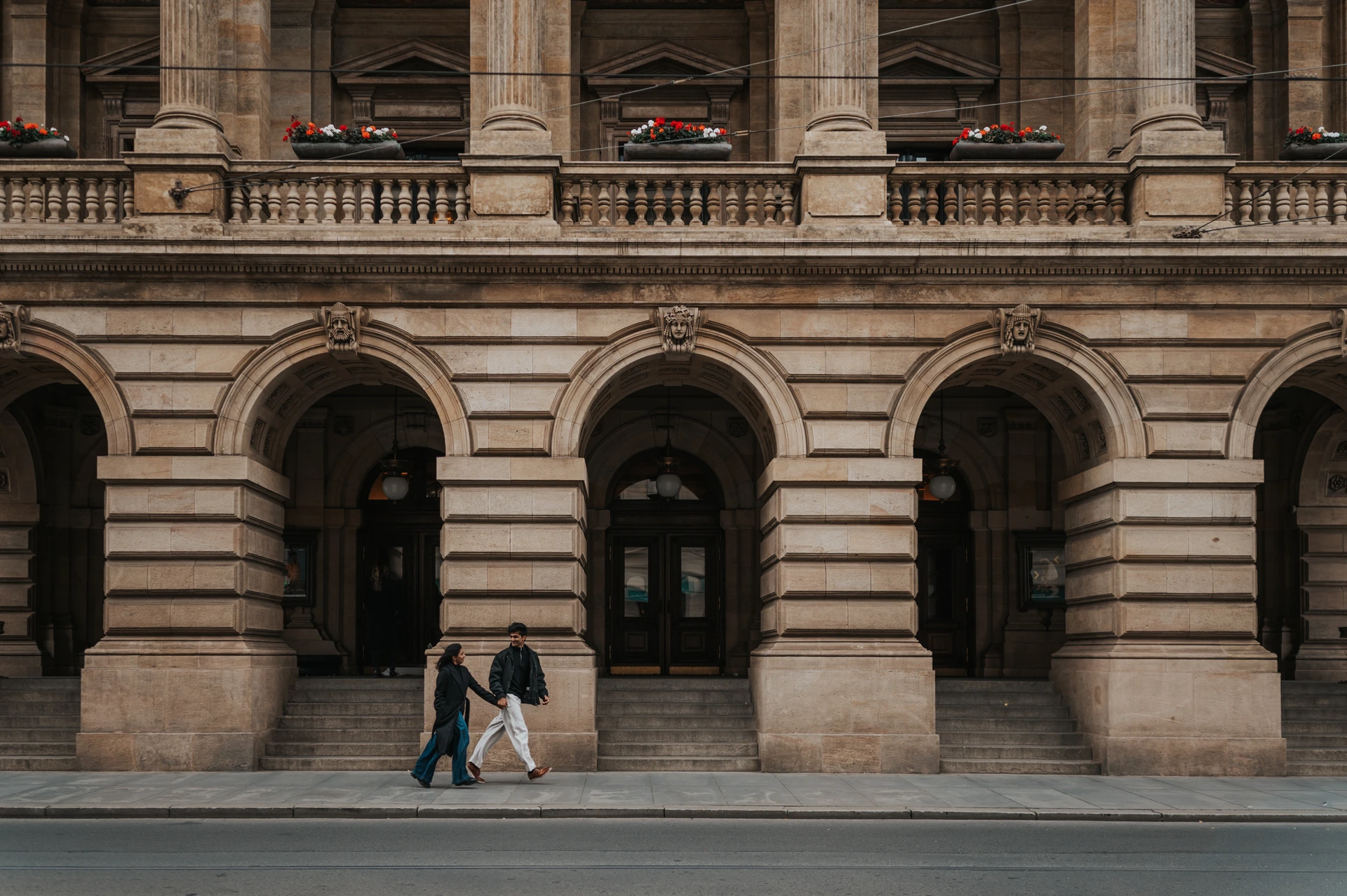 Couple walking hand in hand past a grand stone facade with arches, columns and red flowers in Prague, with tram tracks in the foreground.