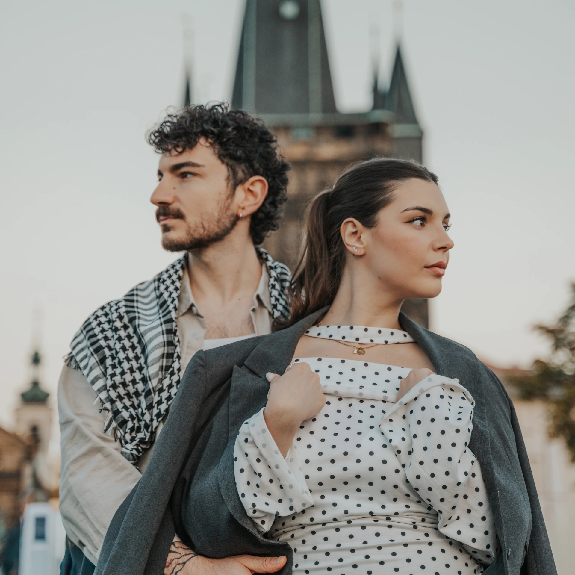 Couple posing close together and looking in opposite directions, with a Charles Bridge tower in Prague behind them.