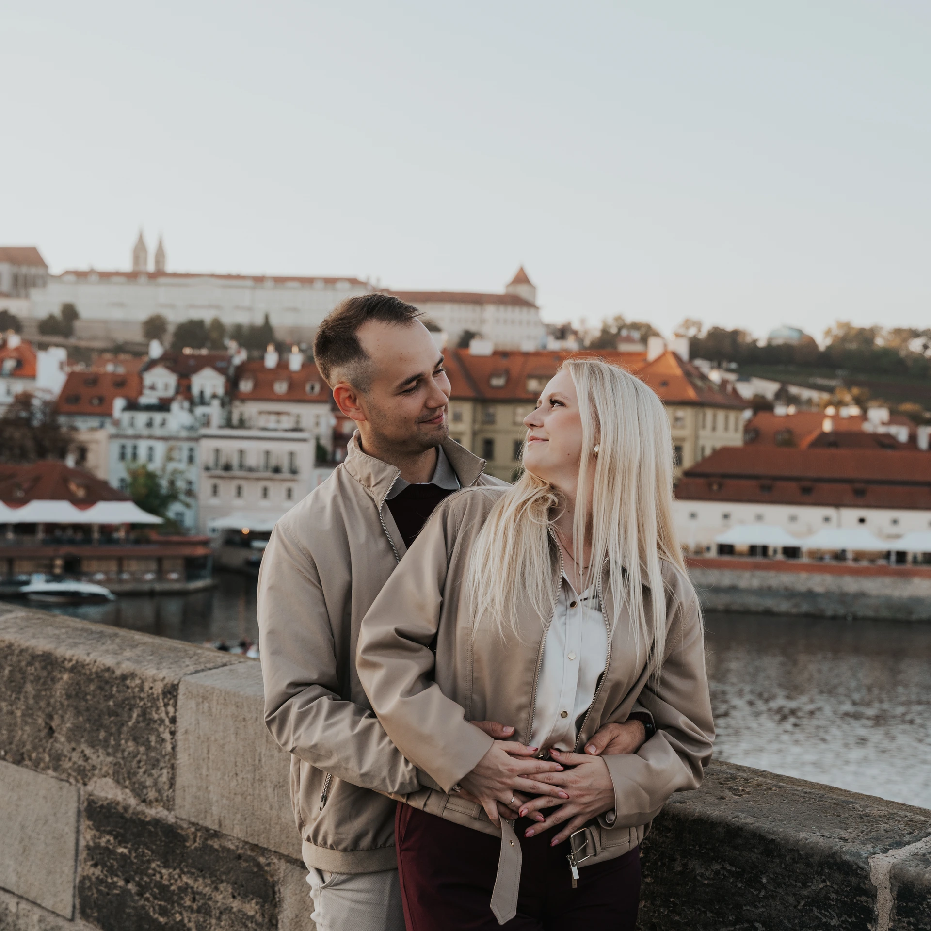 Couple resting their hands on her belly on Charles Bridge, with the Vltava and Prague’s red rooftops in the background.