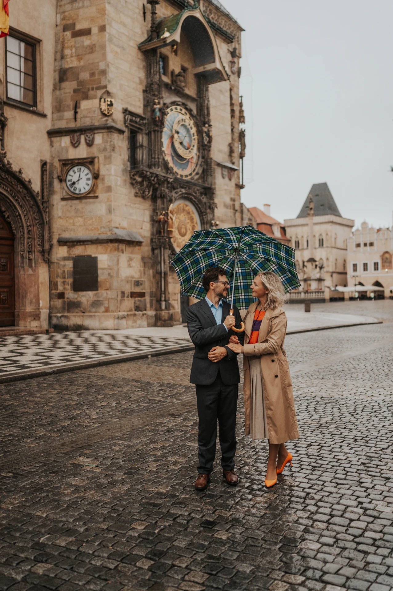 Full-length portrait of the couple sharing an umbrella on wet cobblestones by the Astronomical Clock during their Prague anniversary photoshoot.