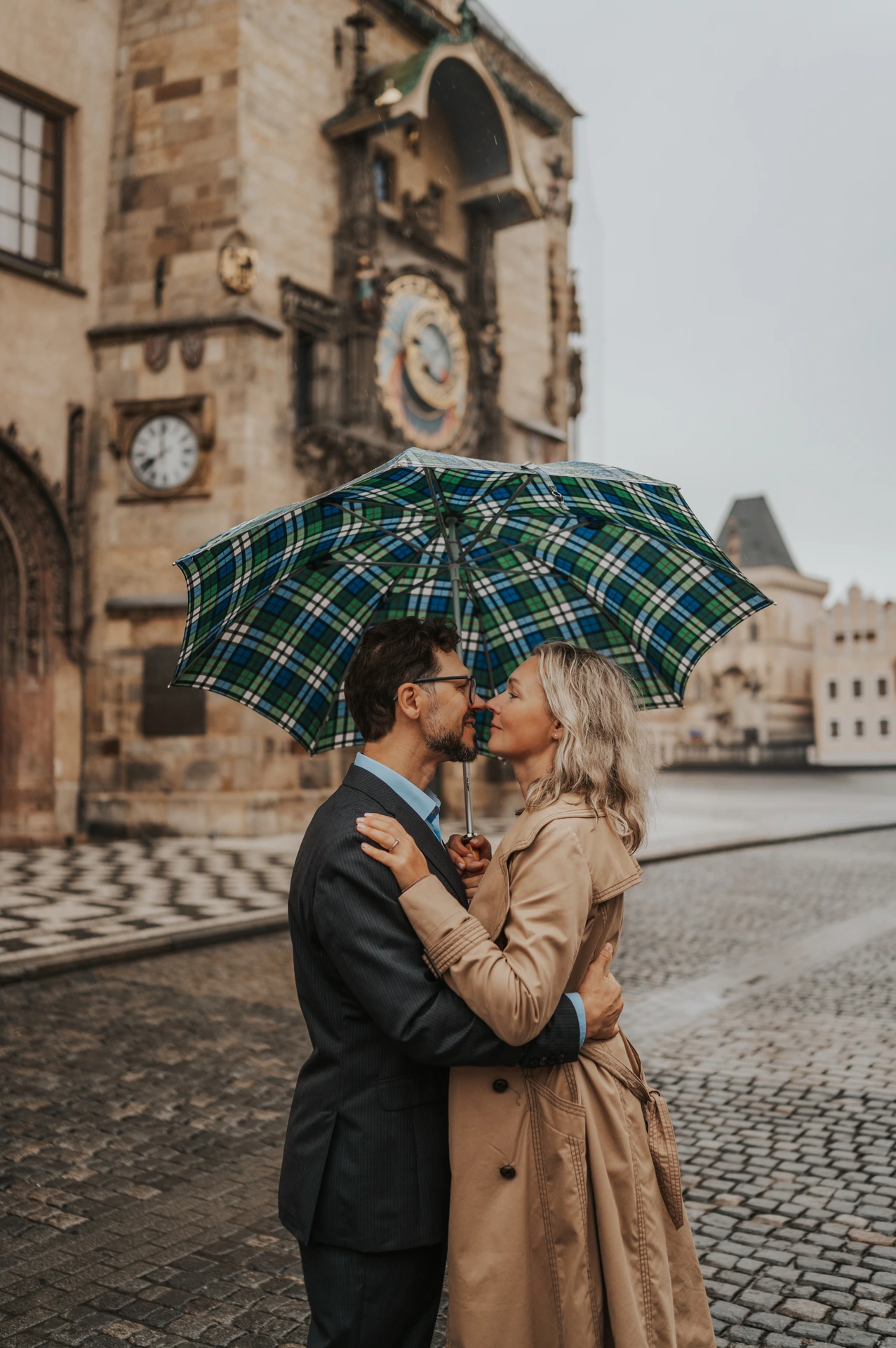Couple kissing beneath a checked umbrella with the Astronomical Clock behind them during their Prague anniversary photoshoot.
