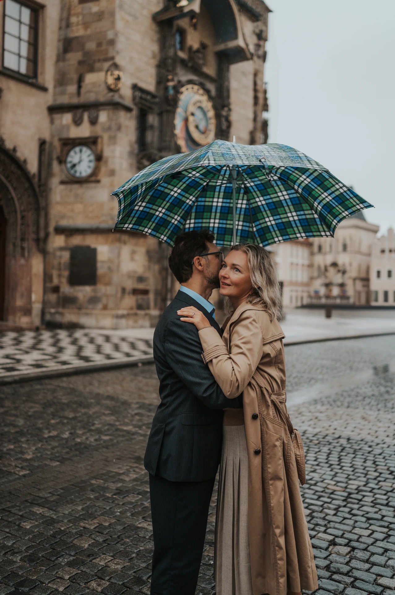 Couple embracing under a checked umbrella as she smiles towards the camera beside the Astronomical Clock on their Prague photoshoot.