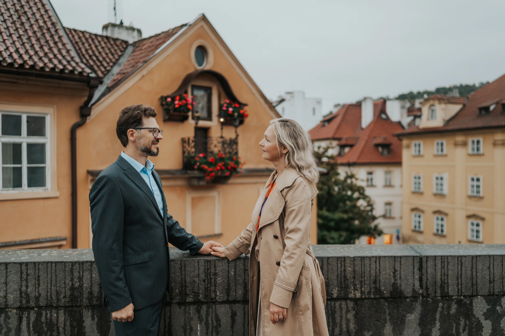 Couple holding hands beside a stone parapet and colourful houses during their anniversary photoshoot in Prague.
