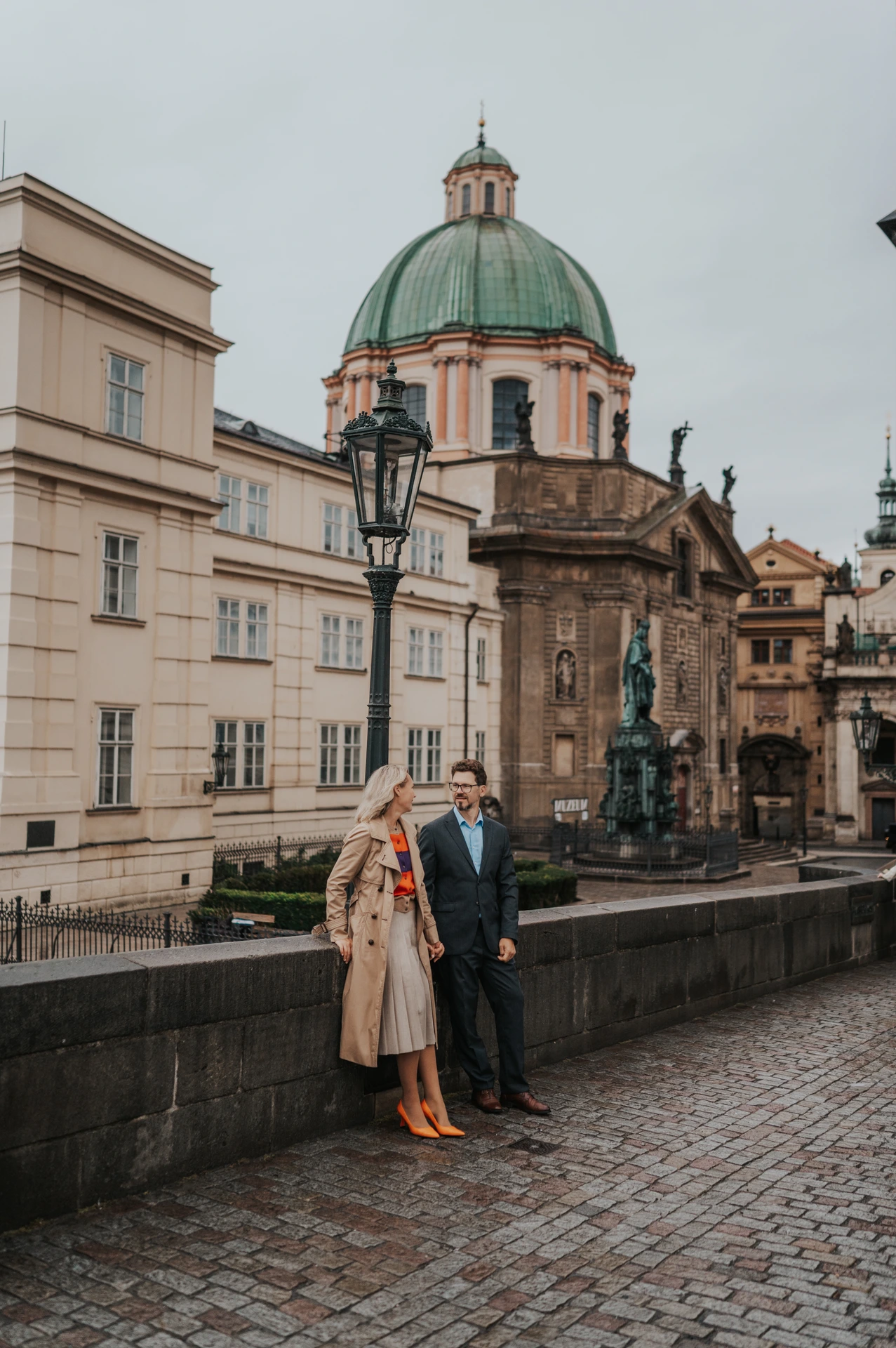 Couple standing by the Charles Bridge wall with a green-domed building behind them during their Prague anniversary photoshoot.