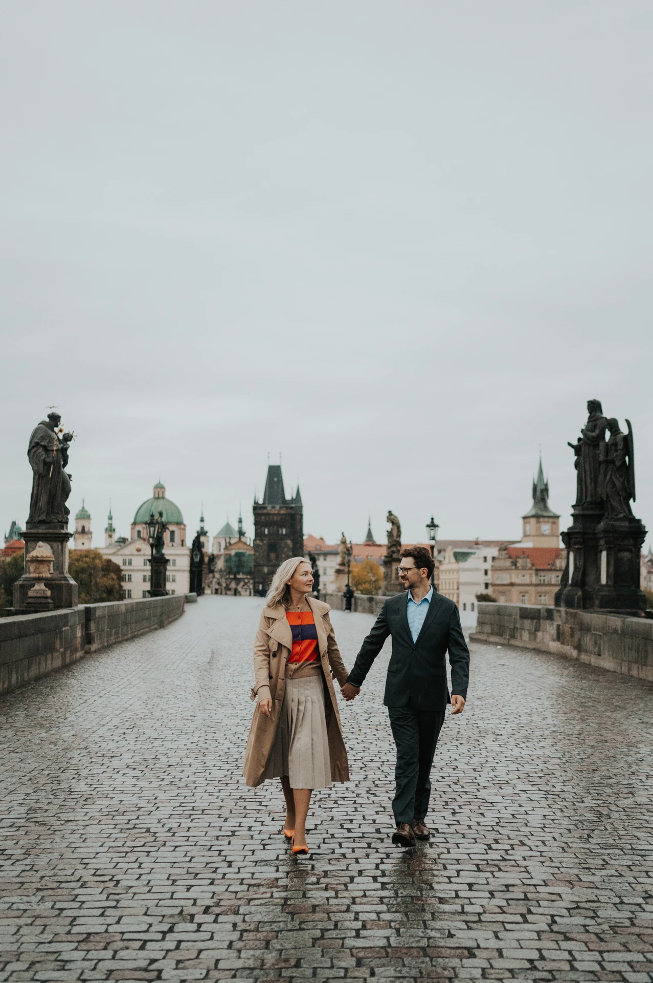 Couple walking hand in hand between the statues of Charles Bridge during a Prague anniversary photoshoot.