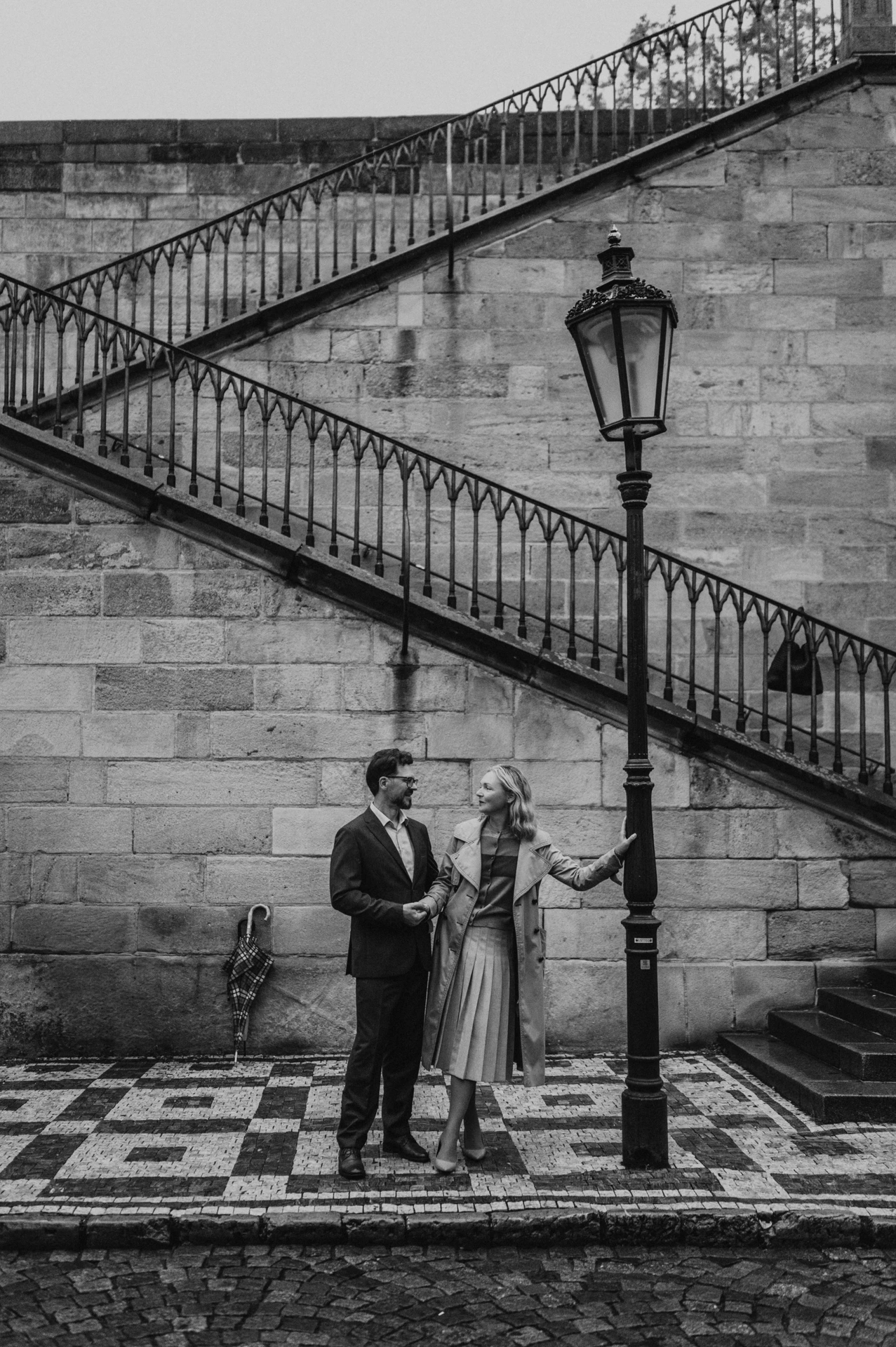 Black-and-white portrait of the couple beside a lamppost and stone staircase on their Prague anniversary photoshoot.