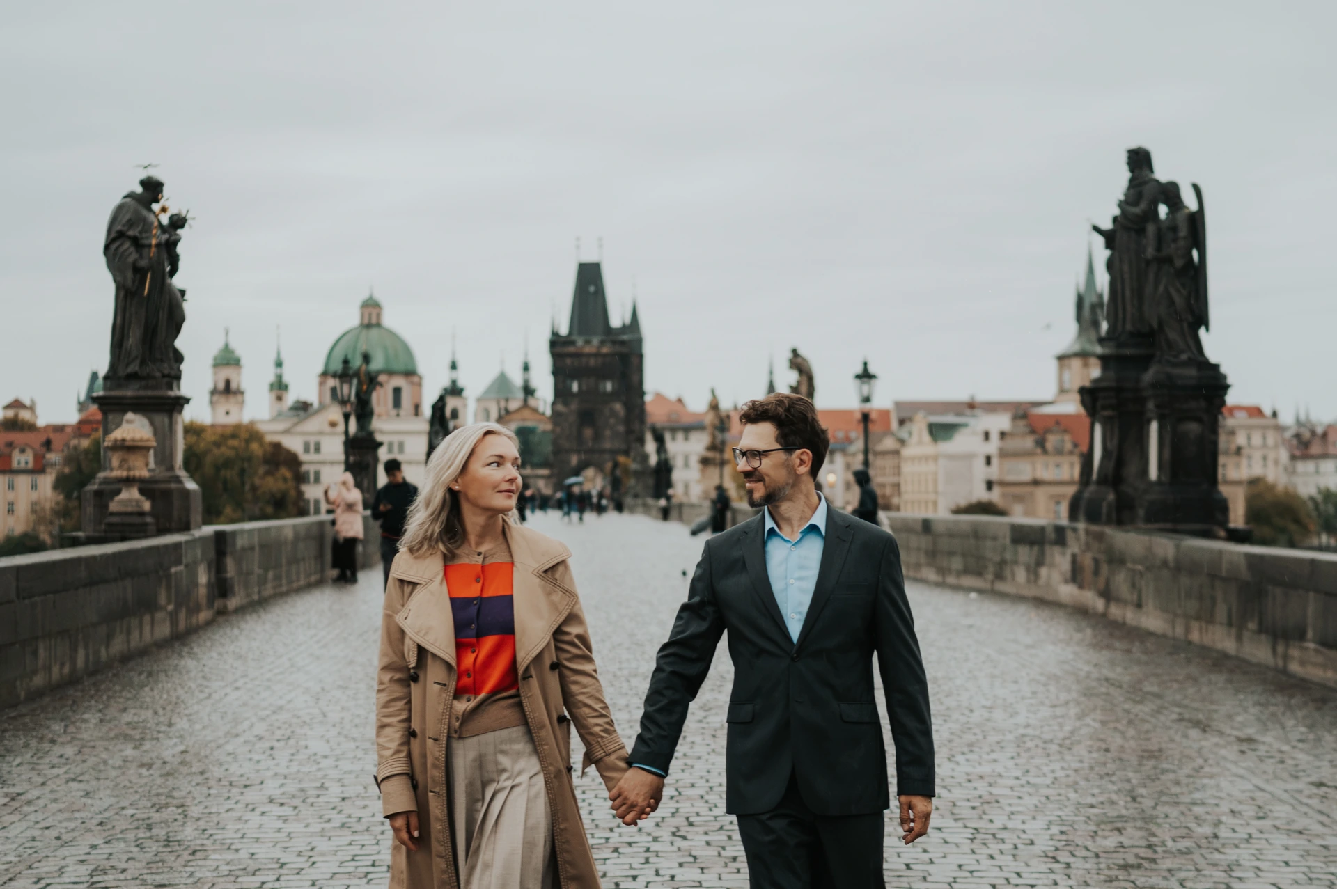 Wide view of the couple walking towards the camera on Charles Bridge during their Prague anniversary photoshoot.