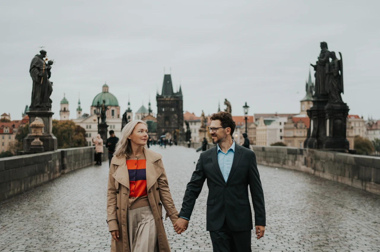 Prague anniversary photoshoot between the Charles Bridge statues Wide view of the couple walking towards the camera on Charles Bridge during their Prague anniversary photoshoot.
