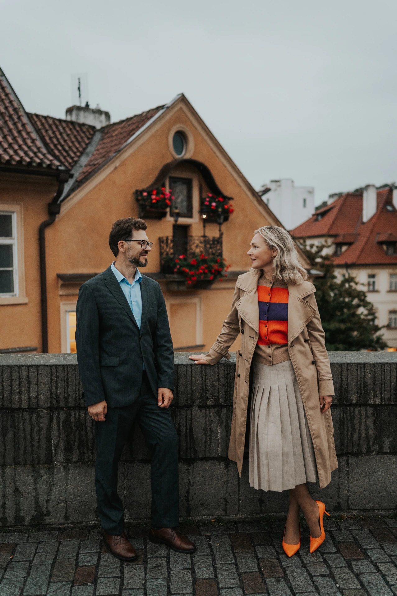 Full-length portrait of the couple holding hands, with her orange heels and a yellow house visible, on their Prague photoshoot.