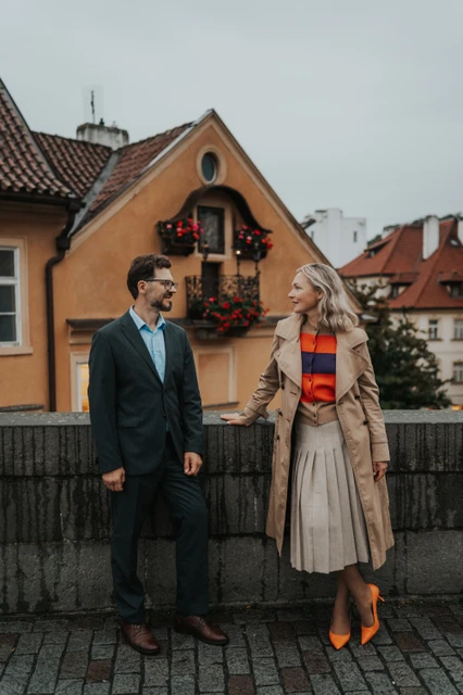 Prague anniversary photoshoot with autumn outfits by Charles Bridge Full-length portrait of the couple holding hands, with her orange heels and a yellow house visible, on their Prague photoshoot.