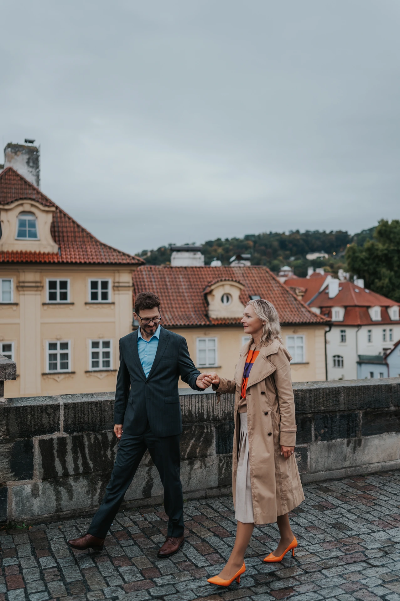 Couple walking hand in hand along the Charles Bridge parapet, with orange shoes against wet paving, on their Prague photoshoot.