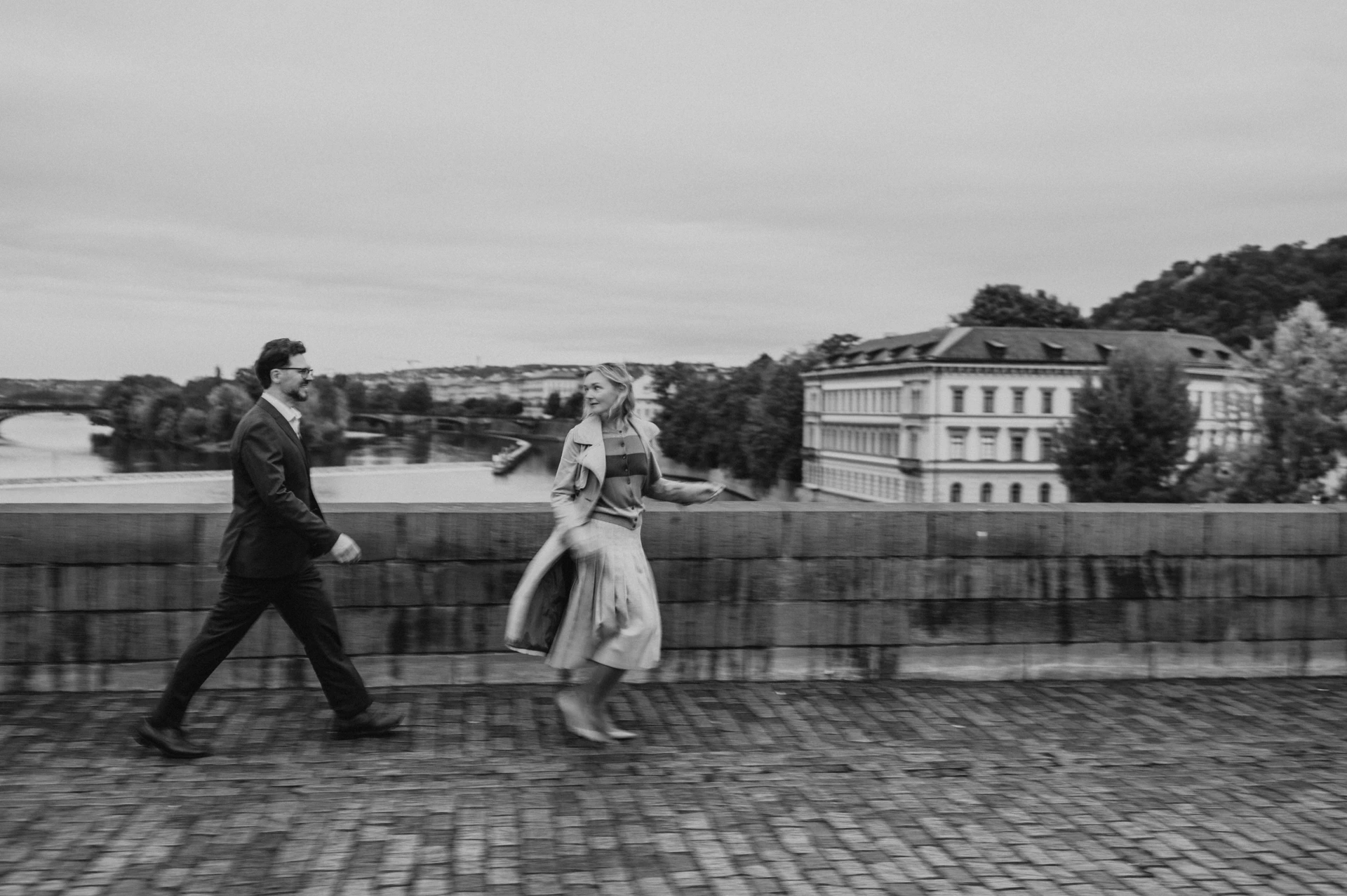 Black-and-white wide photograph of the couple striding hand in hand across Charles Bridge during their Prague anniversary photoshoot.
