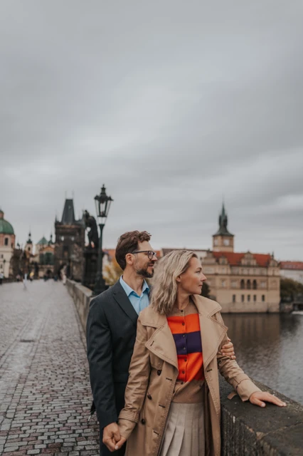 Prague couple photoshoot with a riverside embrace on Charles Bridge Couple looking to the side as he embraces her by the Charles Bridge parapet during their anniversary photoshoot in Prague.