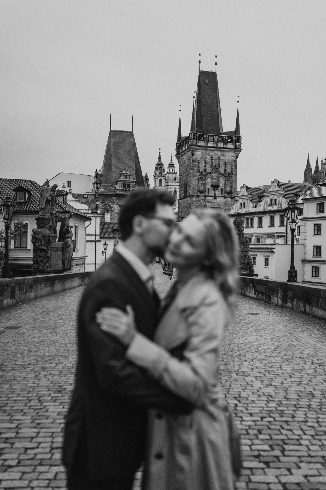 Black-and-white photograph with the kissing couple softly blurred and Charles Bridge towers behind them on their Prague photoshoot.