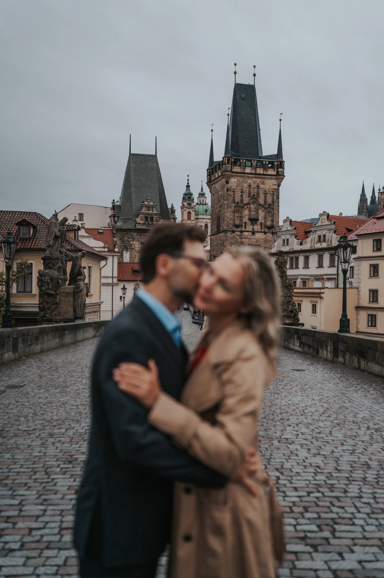 Soft-focus couple kissing in the foreground with the Charles Bridge towers in focus during a Prague anniversary photoshoot.