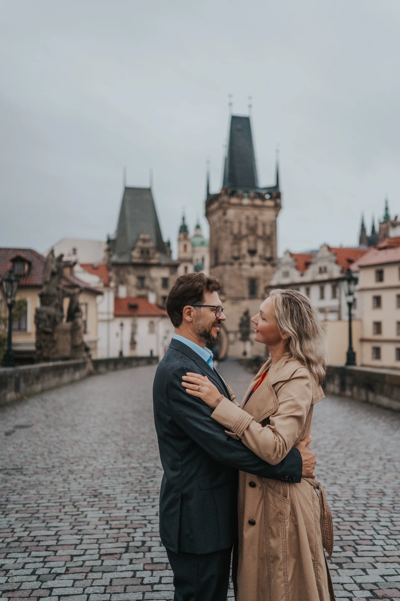 Couple facing each other in an embrace with the Charles Bridge towers behind them during their Prague anniversary photoshoot.