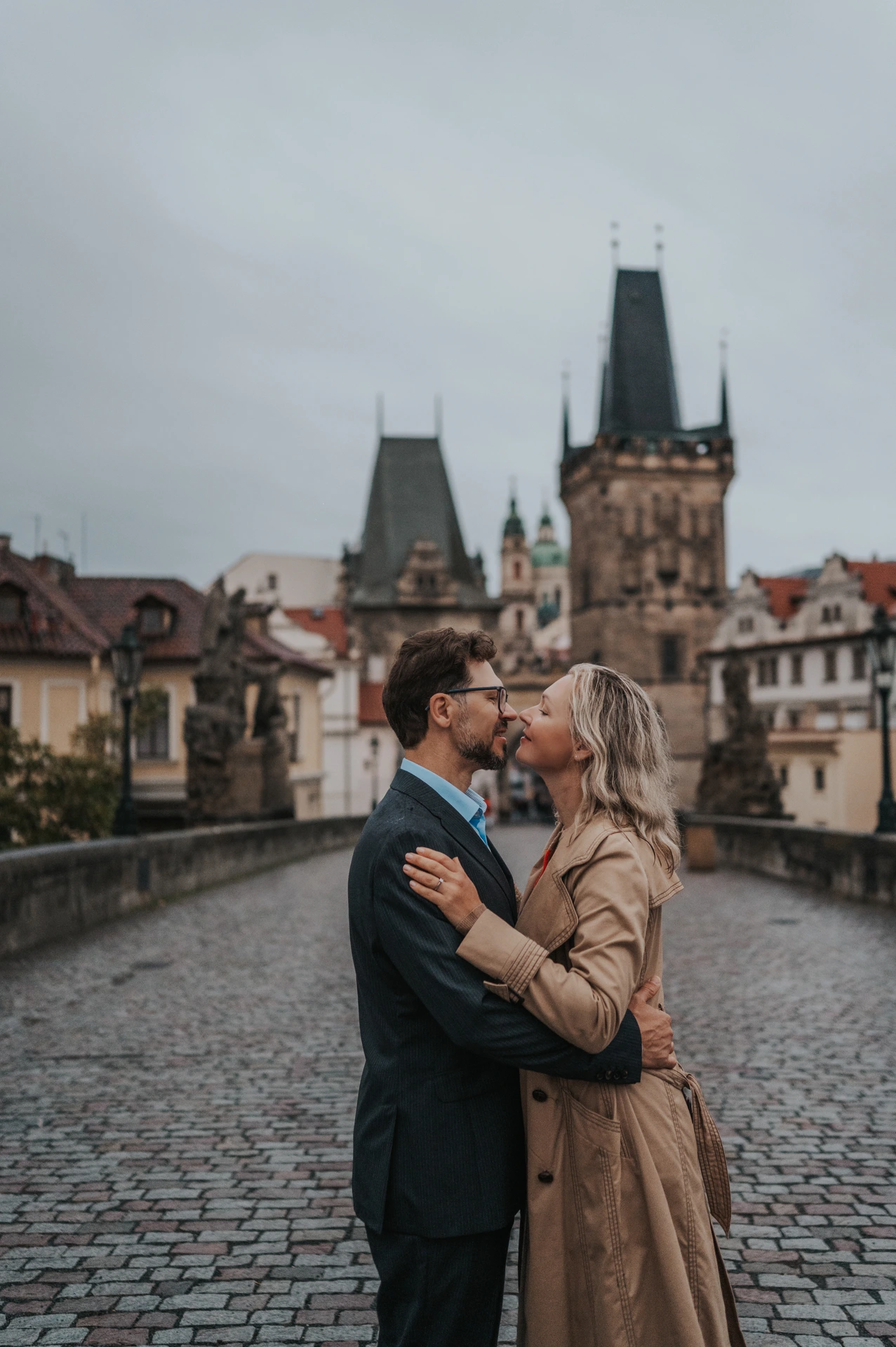 Couple kissing on Charles Bridge beneath a grey autumn sky during their anniversary photoshoot in Prague.