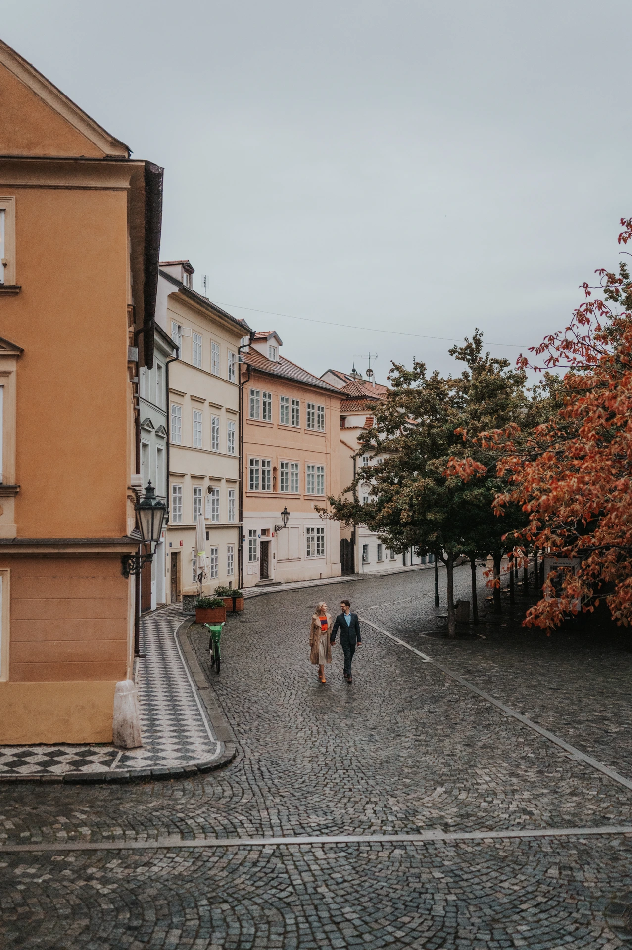 High-angle view of the couple walking across Kampa cobblestones beside an autumn tree during their Prague anniversary photoshoot.