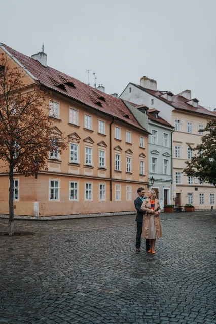 Prague couple photoshoot with an embrace in Kampa Wide portrait of the couple embracing on a cobbled Kampa square beside an autumn tree during their Prague photoshoot.