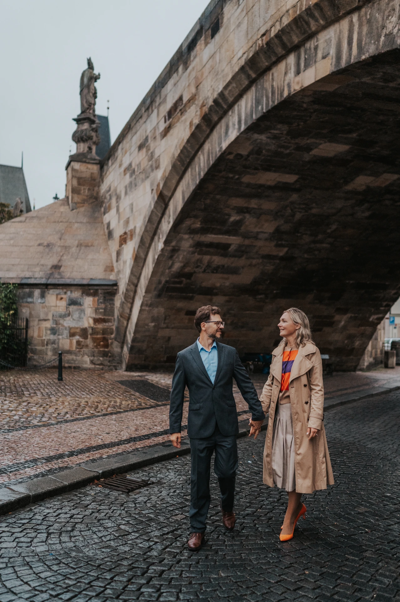 Couple walking hand in hand beneath a Charles Bridge arch on the Kampa side during their Prague anniversary photoshoot.