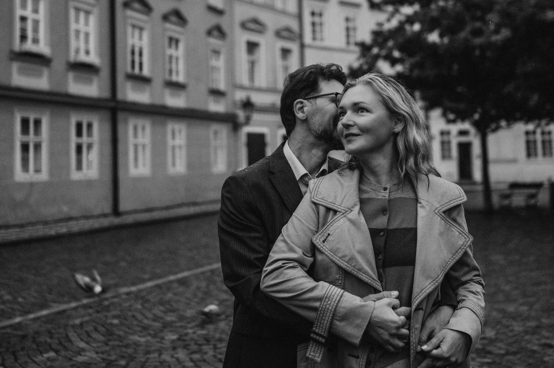 Black-and-white landscape portrait of a cheek kiss and a smile in Kampa during the couple's Prague anniversary photoshoot.