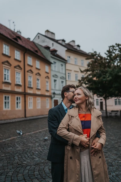 Prague couple photoshoot with a cheek kiss in Kampa Man kissing his wife's cheek as she smiles in front of Kampa houses during their Prague anniversary photoshoot.