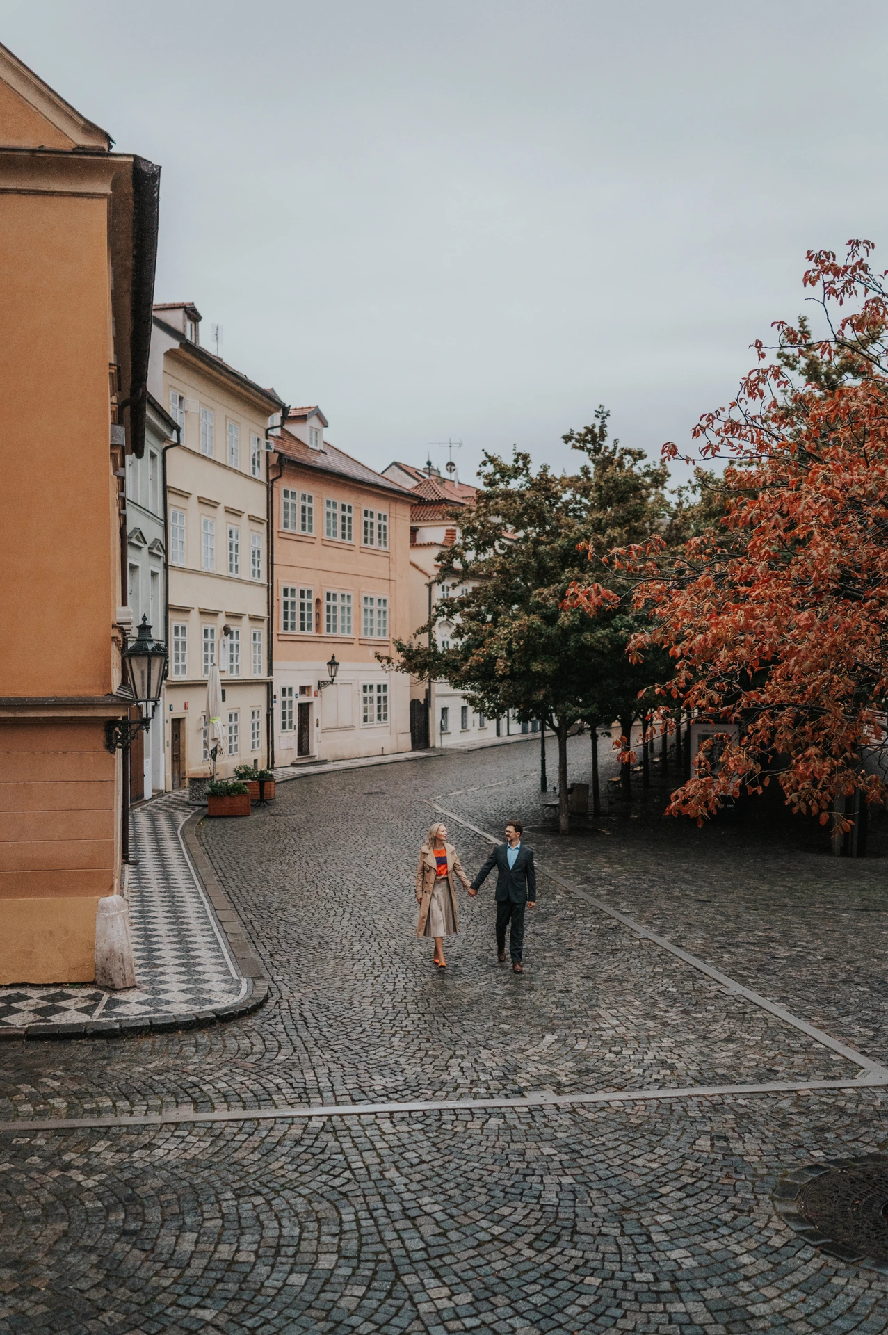Couple holding hands on a cobbled Kampa street below colourful houses during their anniversary photoshoot in Prague.