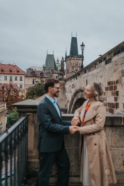 Prague couple photoshoot by the Kampa steps with bridge towers behind Soft-focus couple holding hands beside the Kampa stair railing, with Charles Bridge towers behind, on their Prague photoshoot.
