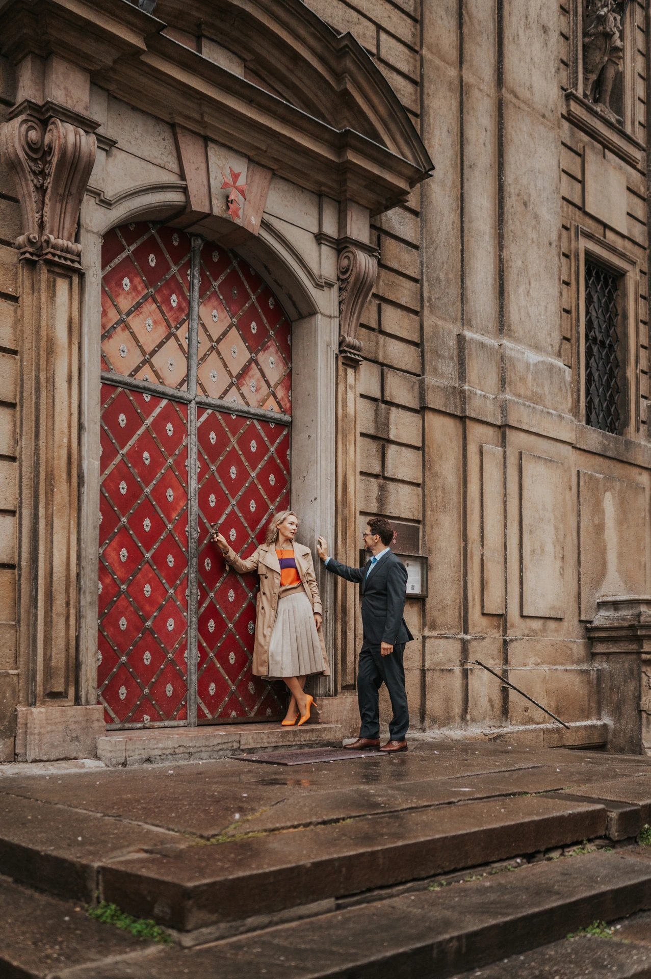 Couple holding hands while stepping away from the red Old Town doorway during their Prague anniversary photoshoot.