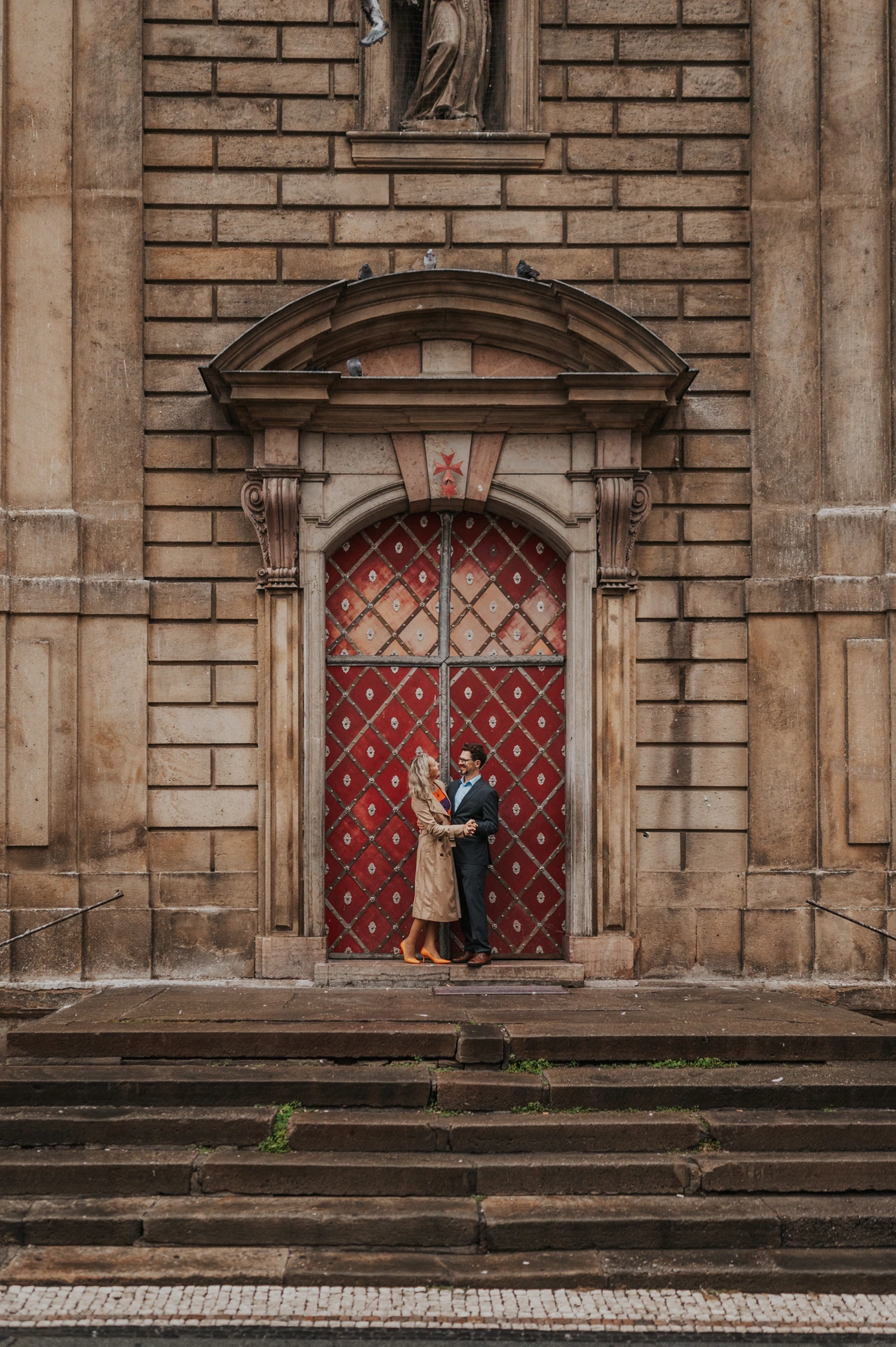 Distant portrait of the couple at a tall patterned red doorway in Old Town during their Prague anniversary photoshoot.