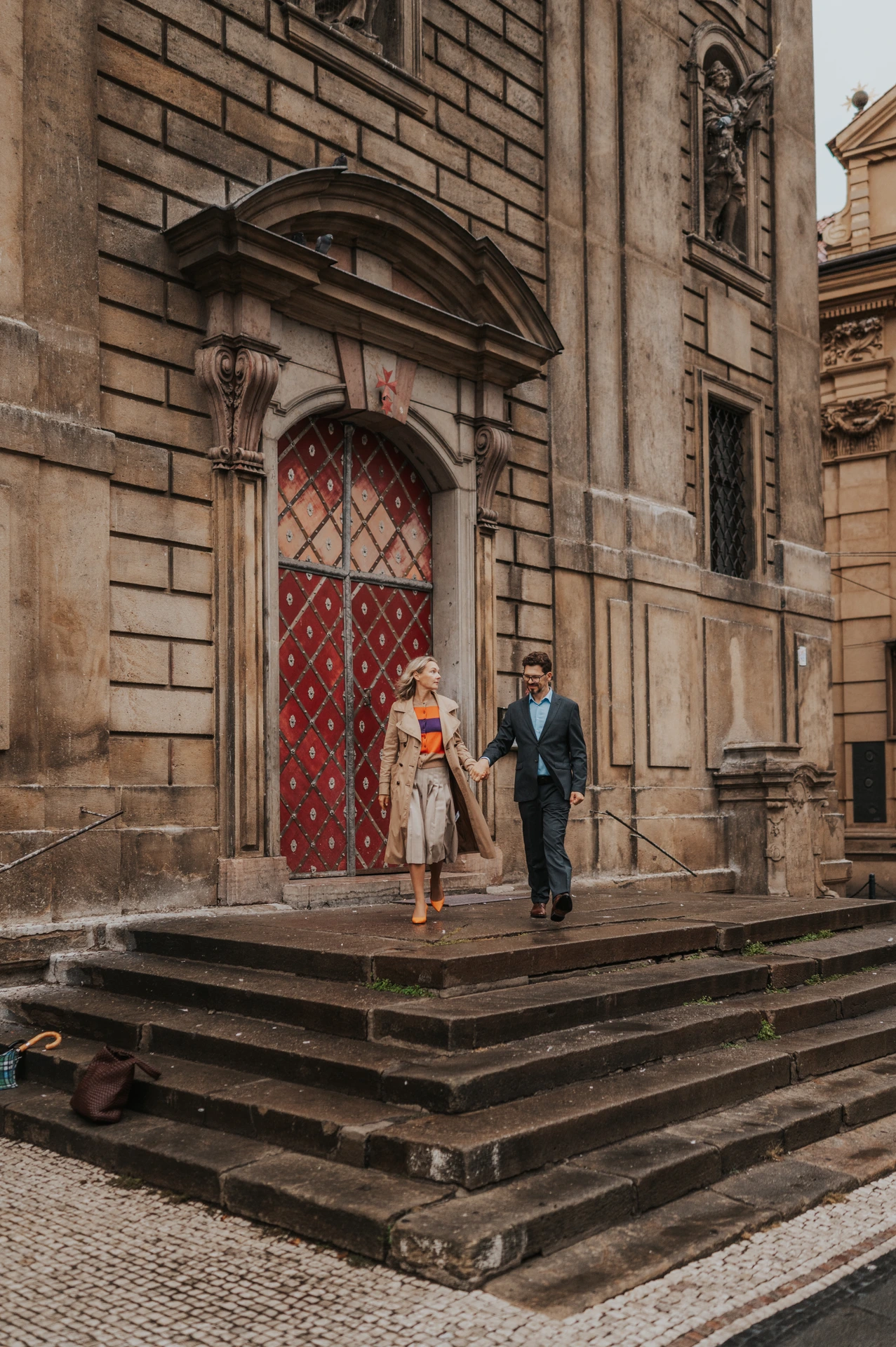 Couple walking down wet stone steps beneath a red doorway in Old Town during their Prague anniversary photoshoot.