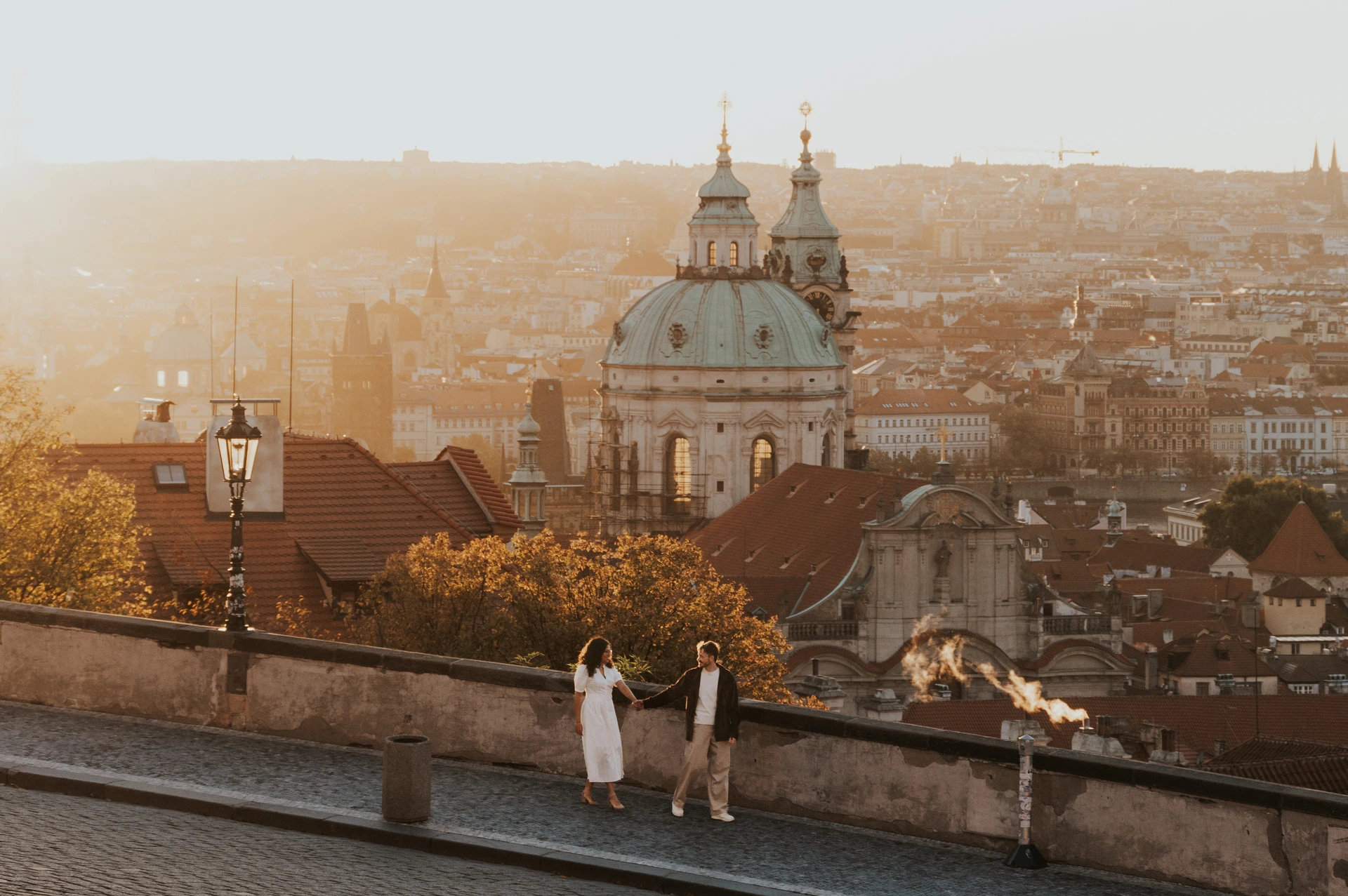 Prague anniversary photoshoot: wide portrait of the couple with a domed church and sunrise behind them. Wide portrait of the couple with a domed church and sunrise behind them during an anniversary couple photoshoot in Prague.