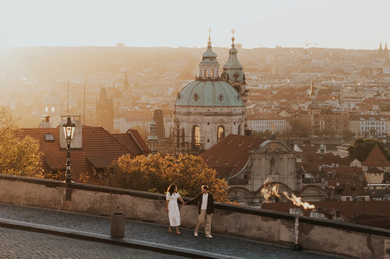 Prague anniversary photoshoot: wide portrait of the couple with a domed church and sunrise behind them. Wide portrait of the couple with a domed church and sunrise behind them during an anniversary couple photoshoot in Prague.